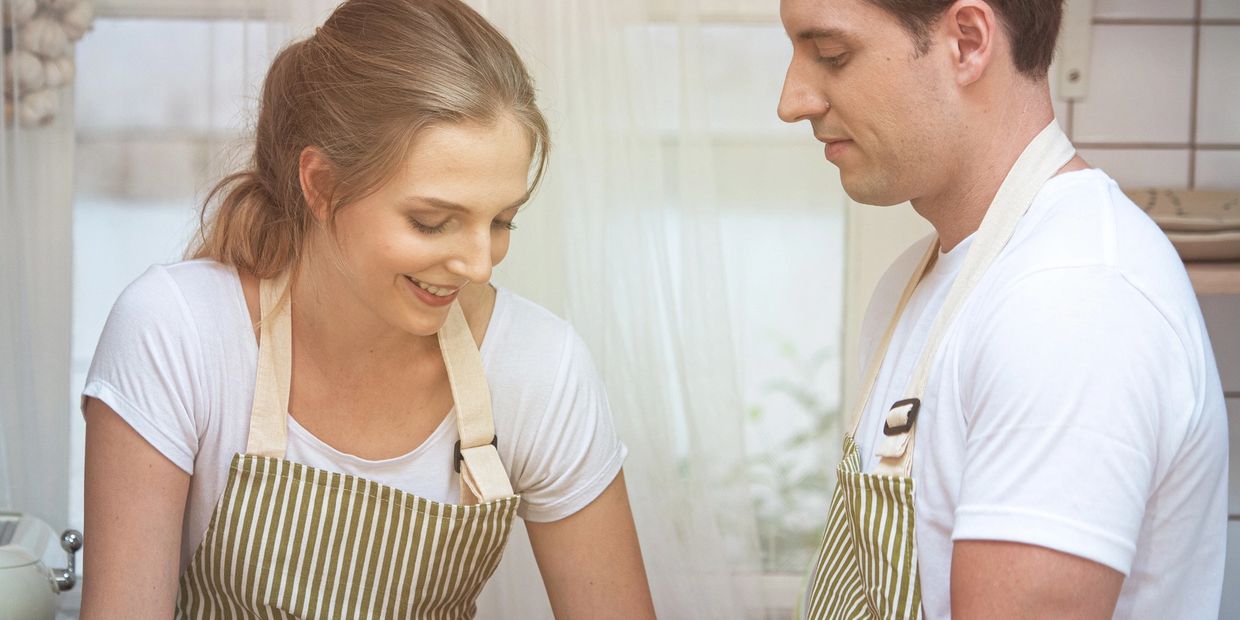 Couple wearing aprons cooking together in a bright kitchen.