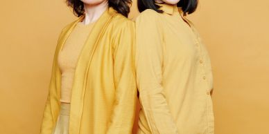Two women wearing yellow clothing standing back to back against a yellow background.