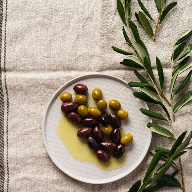 Several varieties of fresh olives in different ceramic plates on an old vintage gray napkin tablecloth table background. Natural product concept. Rustic vintage set of cutlery. Top view, copy space.