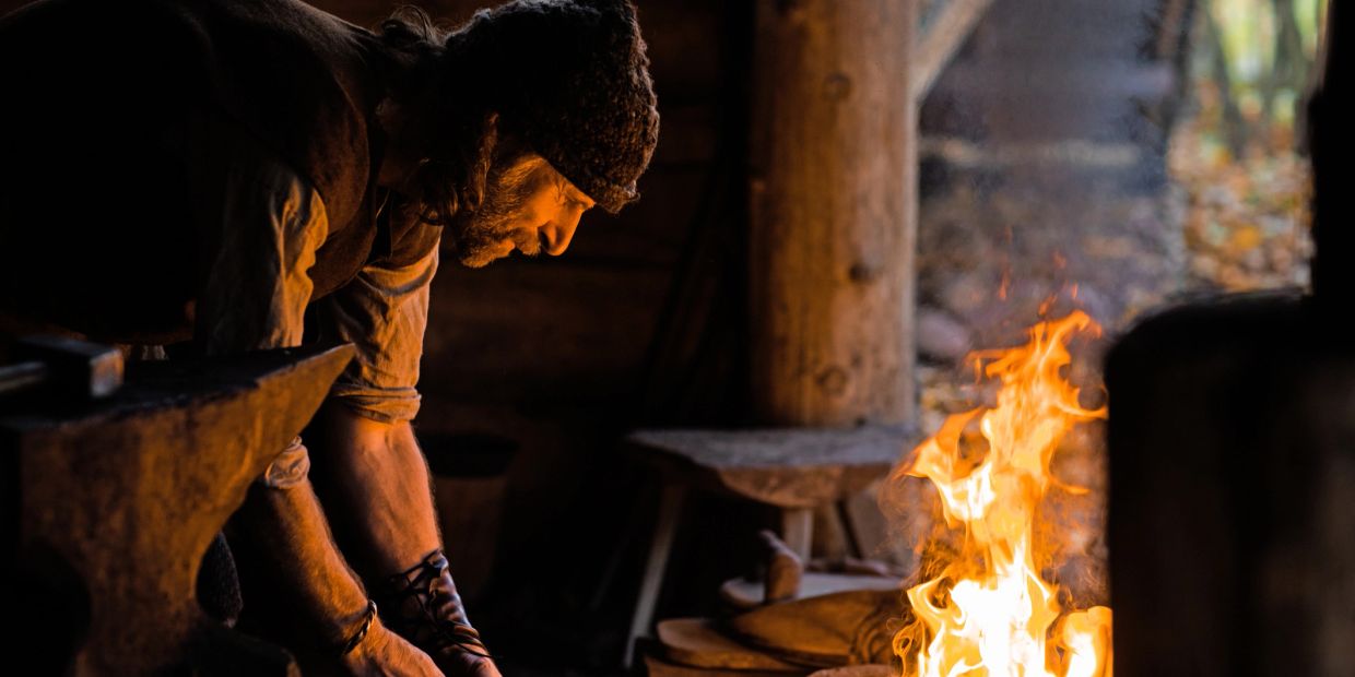 Blacksmith working near fire in dim workshop.