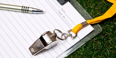 A silver whistle with a yellow lanyard on a clipboard with lined paper and a pen.
