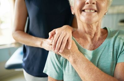 Elderly woman smiling with a hand on her shoulder.