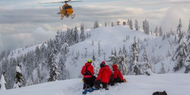 Rescue team in red gear watches a yellow helicopter over snowy mountains.
