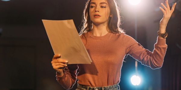 Young woman passionately rehearsing a script on stage under bright lights.