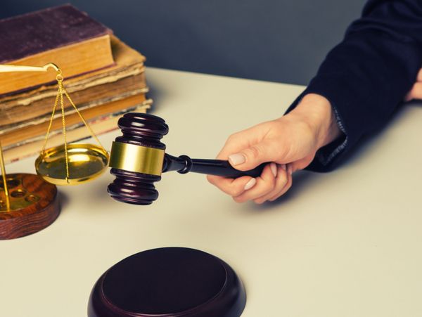 A hand holding a judge's gavel near a sound block and scales of justice.