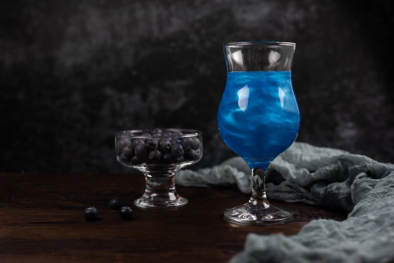Sparkling glitter blue drink. A cocktail in a glass goblet stands on a wooden table with a transparent bowl of blueberries. Festive still life with copy space on a dark background.
