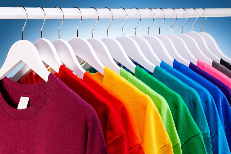 row of many fresh new fabric cotton t-shirts in colorful rainbow colors hangng on clothes rail in wrdrobe. Various colored shirts on blue white background. diy printing fashion concept.