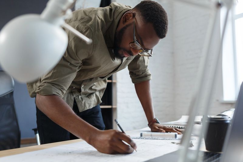 Afro-American architect working in office with blueprints.Engineer inspect architectural plan, sketching a construction project. Portrait of black handsome man sitting at workplace. Business concept.