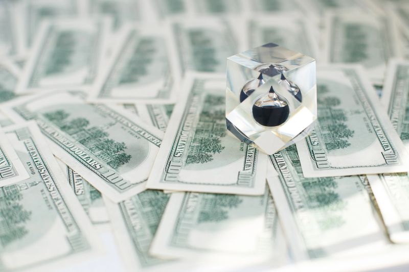 Drop of black oil of Brent brand in crystal cube laying on the background of hundred dollars bills. Crisis in global financial market, falling and rising oil prices, quotes, changes in exchange rate.