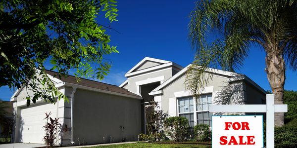 A single-story house with a "For Sale" sign in the front yard under a clear blue sky.