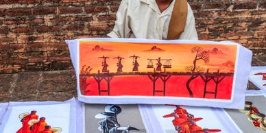 A person displays vibrant traditional artwork featuring silhouetted figures against a sunset.