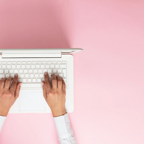Hands typing on a white laptop keyboard against a pink background.