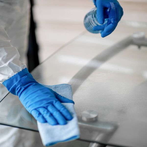 Person in protective suit cleaning a glass table with spray and cloth.