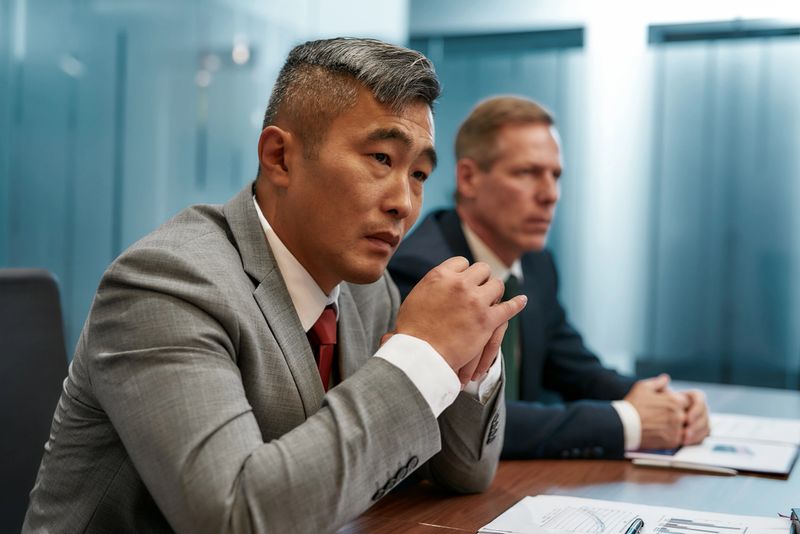 Portrait of focused asian businessman in formal wear listening carefully while sitting with his caucasian collegue at office desk in the modern office. Business concept. Partnership. Office life