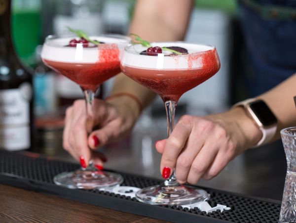 Two red cocktails being placed on a bar by a person with red nail polish.