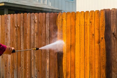 A person pressure washing a wooden fence, revealing its bright orange color.