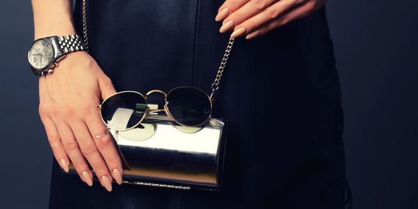 Woman holding a shiny silver clutch and trendy sunglasses against a black outfit.