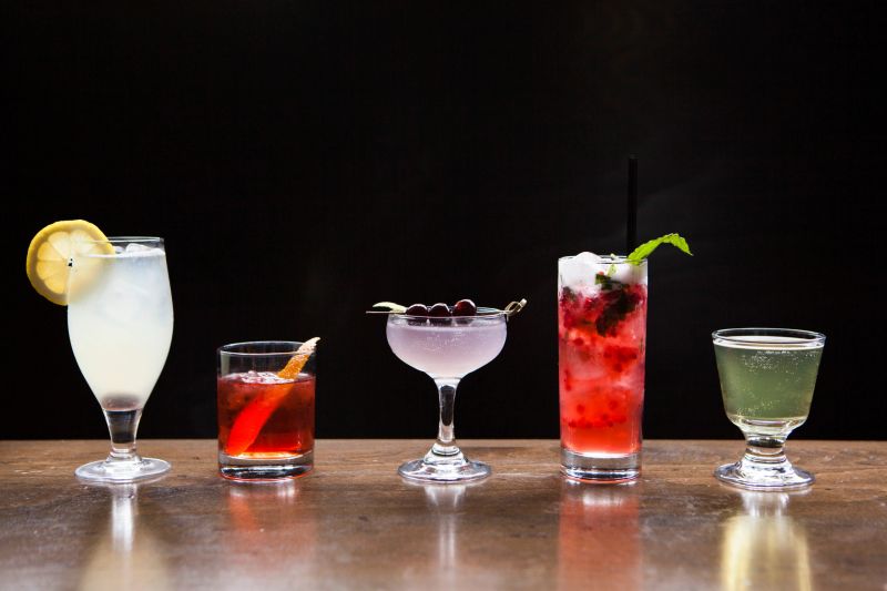 Five cocktails of different heights, shapes, and colors (red, green, purple, yellow) on a wooden table with a black background.