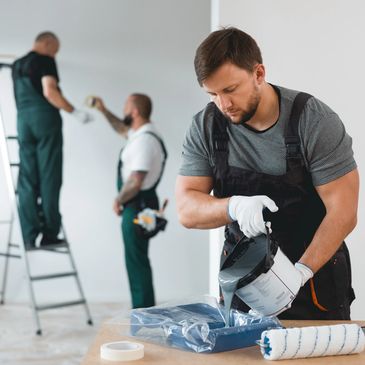 Painter pouring gray paint into a tray while two others prepare a wall.