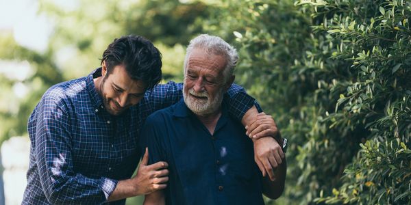A young man affectionately supporting an elderly man outdoors.