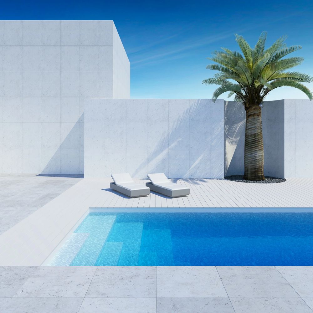 Minimalist poolside with two white loungers and a palm tree against a clear blue sky.