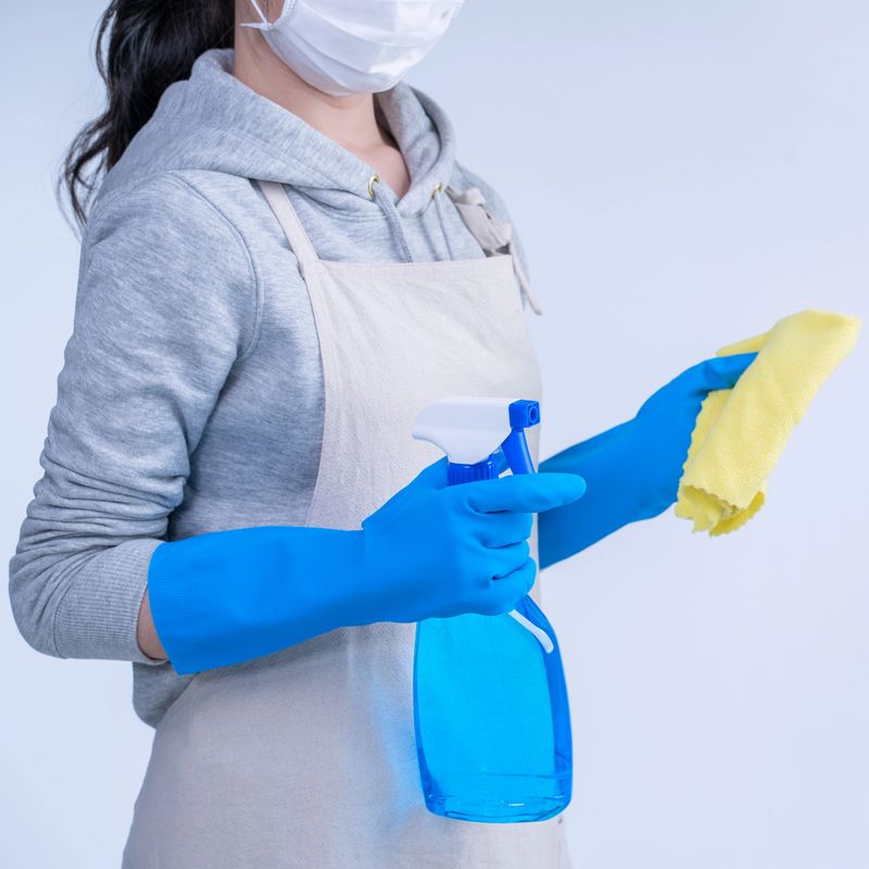 Young woman housekeeper in apron is doing cleaning with blue gloves, wet yellow rag, spraying bottle cleaner, close up, copy space, blank design concept.