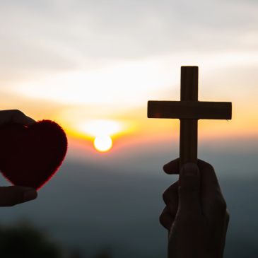 Hands holding a heart and a wooden cross against a sunset.