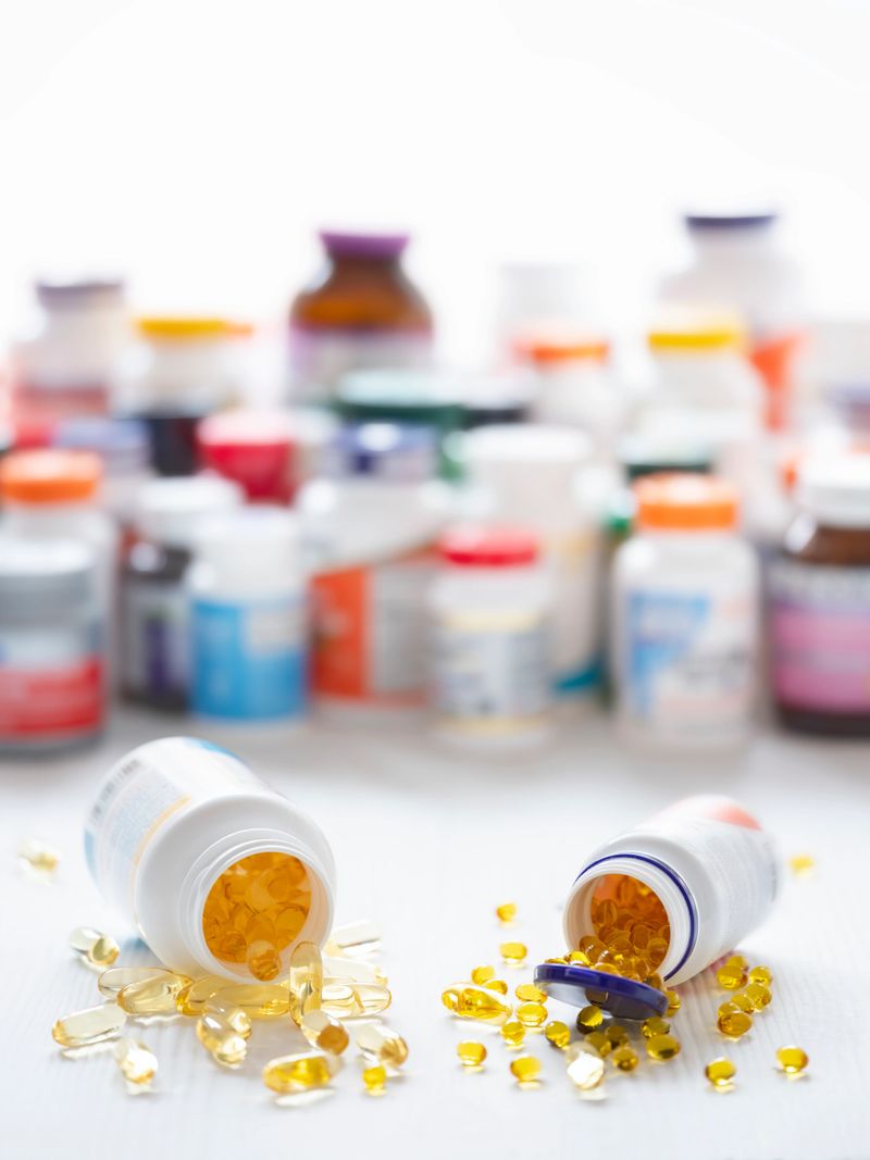 Colorful display of many types of nutritional supplements, with two horizontal bottles in focus spilling gelatin capsules out onto the table, in the foreground. Scene set on a wood table background. Very shallow depth of field, focus on the gelatin capsules in the foreground.