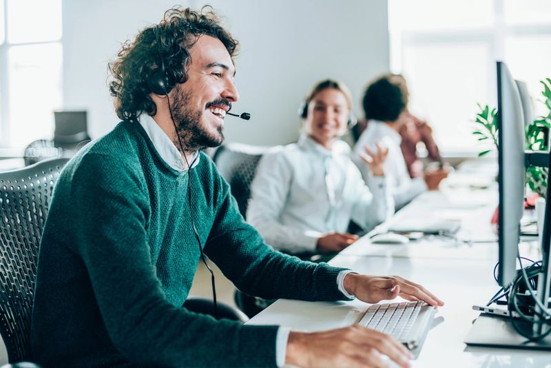 Smiling handsome young businessman working in call center. Shot of a cheerful young man working in a call center with his team. Confident male operator is working with colleagues. Call center operators sitting in a row at desks.