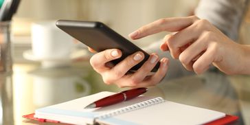Person using smartphone with notebook and pen on table.