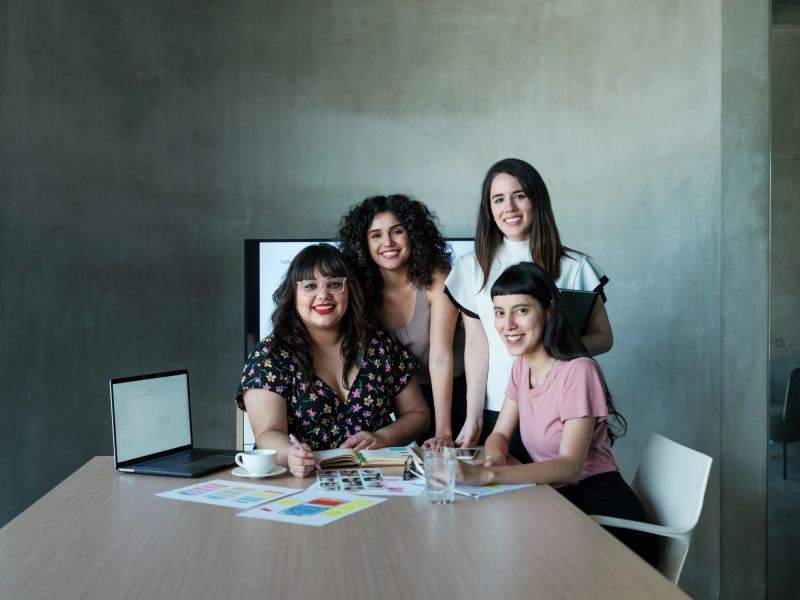 Beautiful young lating businesswomen sitting and standing together at a table at work, looking at the camera and smiling.