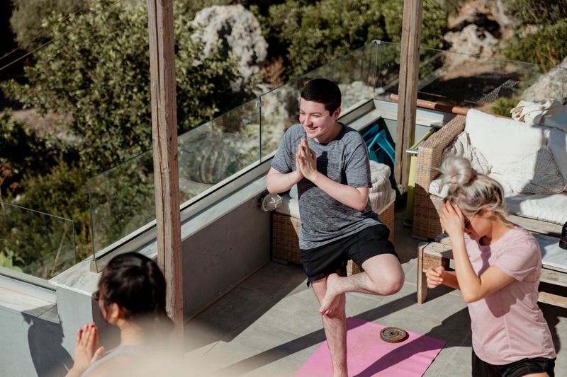 A group of young people are taking an outdoor yoga class in the sun. They are stretching on yoga mats and looking positive.