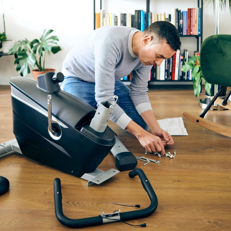 a caucasian man assembling the different pieces of a stationary bicycle in the living room of his house