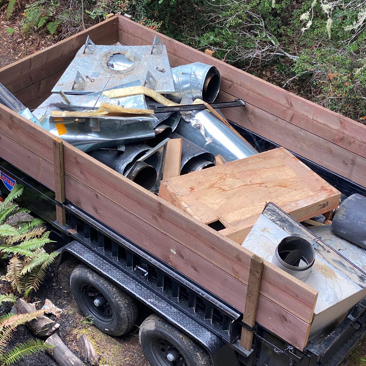 Trailer loaded with metal ducts and wooden planks in a forested area.