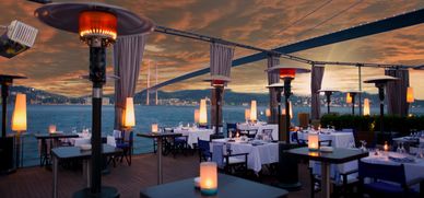 Elegant outdoor dining area with heaters overlooking a bridge at sunset.