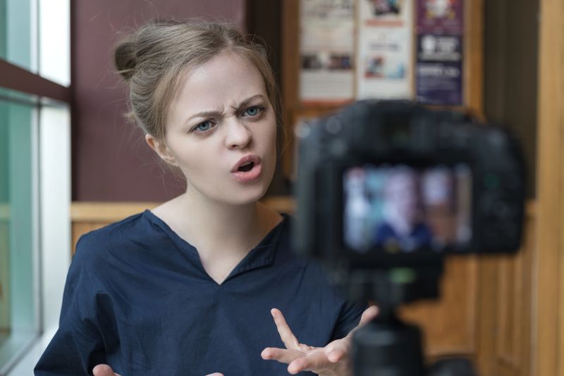 Smiling young caucasian girl woman making a video blog (vlog) with camera in a cafe,