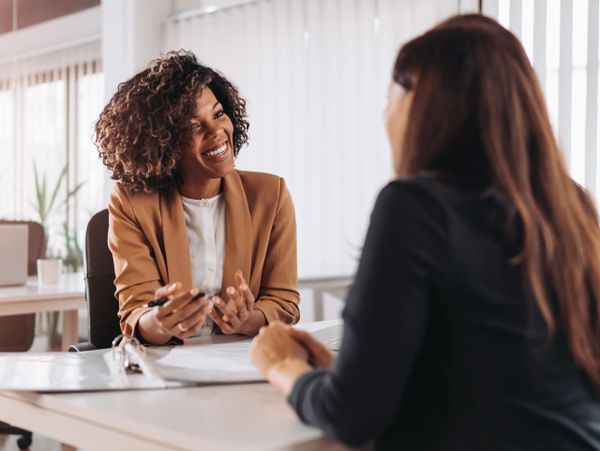 Two women engaged in a friendly office conversation across a desk.