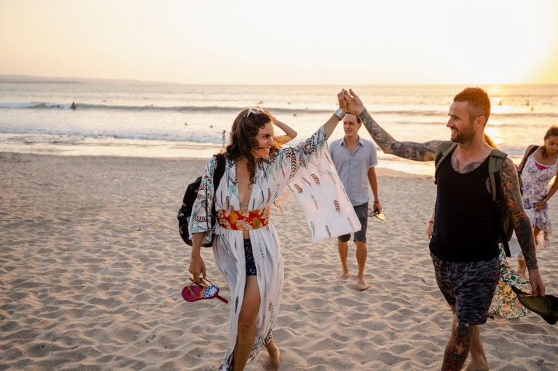 A group of male and female friends walking on Kuta beach in Bali, Indonesia. The main focus is a heterosexual couple holding hands in the air while looking at each other.