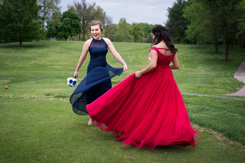 Blond teenager in a formal navy long length dress dances outdoors on a grassy lawn with her best friend wearing a red chiffon dress on a spring day before prom