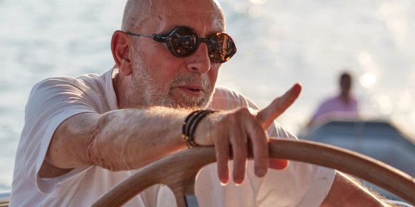 Older man steering a boat on calm water, wearing sunglasses.