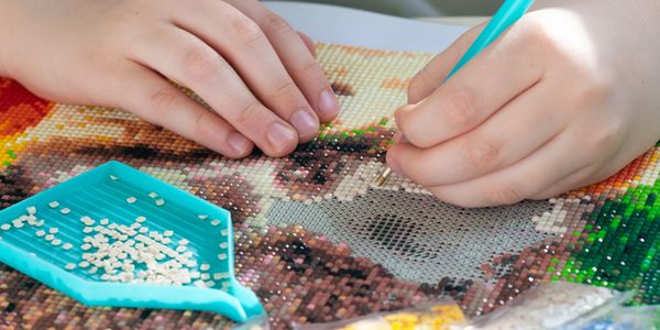 Hands placing beads on a diamond painting canvas with a tool and tray.
