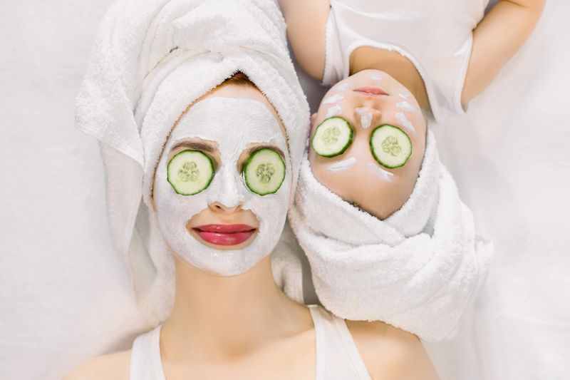 Mother and little daughter having spa procedures together. They are in white bath towels on head and with slices of cucumber on their eyes. Woman has white facial mask on her skin.