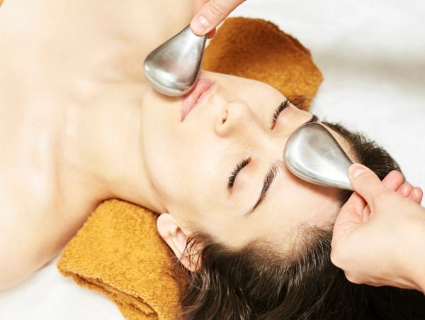 Woman receiving a facial massage with metal tools while lying on a towel.