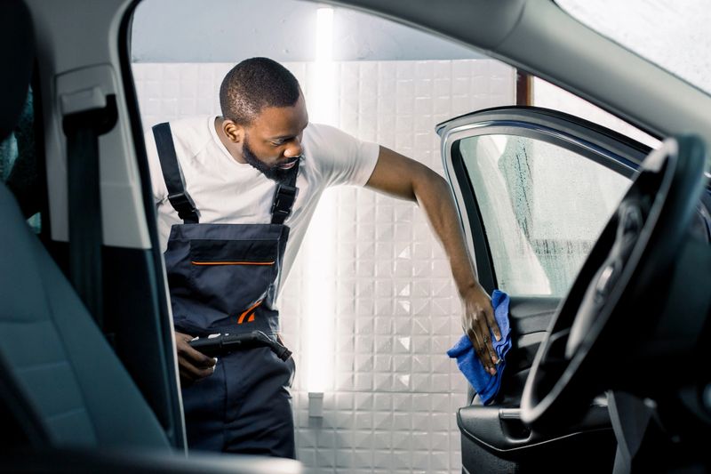 Young African employee of car-care center in overalls and t-shirt holding steam generator and microfiber cloth for cleaning of salon of the car. Car wash and detailing concept.