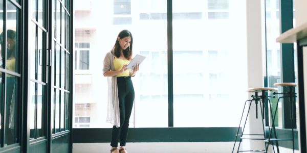 Woman standing by large windows, using a tablet in a bright, modern space.