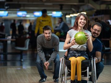 A woman in a wheelchair holding a bowling ball, smiling with friends at a bowling alley.