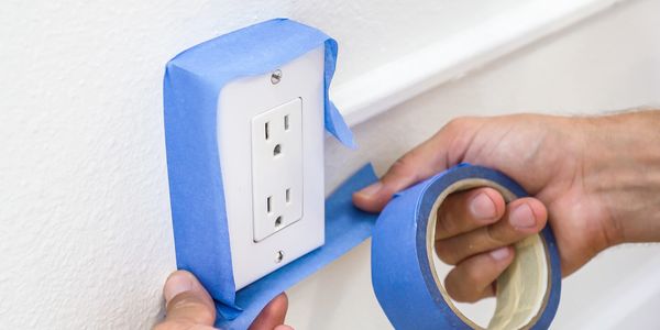 Hands applying blue painter's tape around an electrical outlet on a white wall.