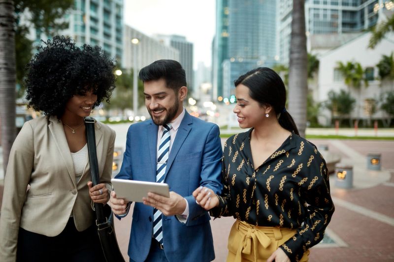 Front view of three Hispanic and African-American business associates standing outdoors in downtown Miami and looking at data on digital tablet.