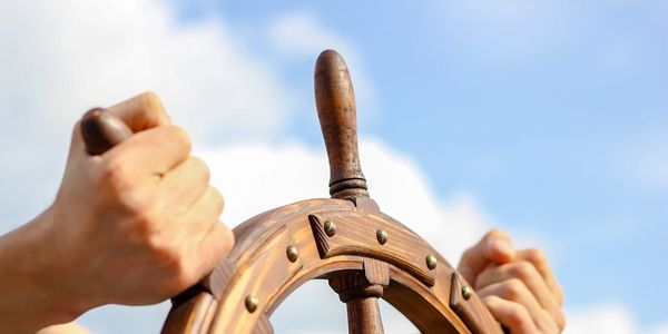 Hands gripping a wooden ship steering wheel against a blue sky.