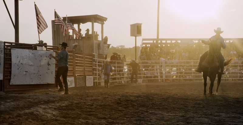 A Cowboy on a Horse Rides through a Stadium Full of People at a Bull Riding/Rodeo Event as another Cowboy Attaches a Covering to a Bull's Pen at Sunset